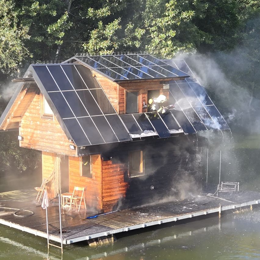 Ein kleines Holzhaus auf einem See mit Solarpanelen auf dem Dach steht in Flammen, und Feuerwehrleute versuchen, das Feuer zu löschen.