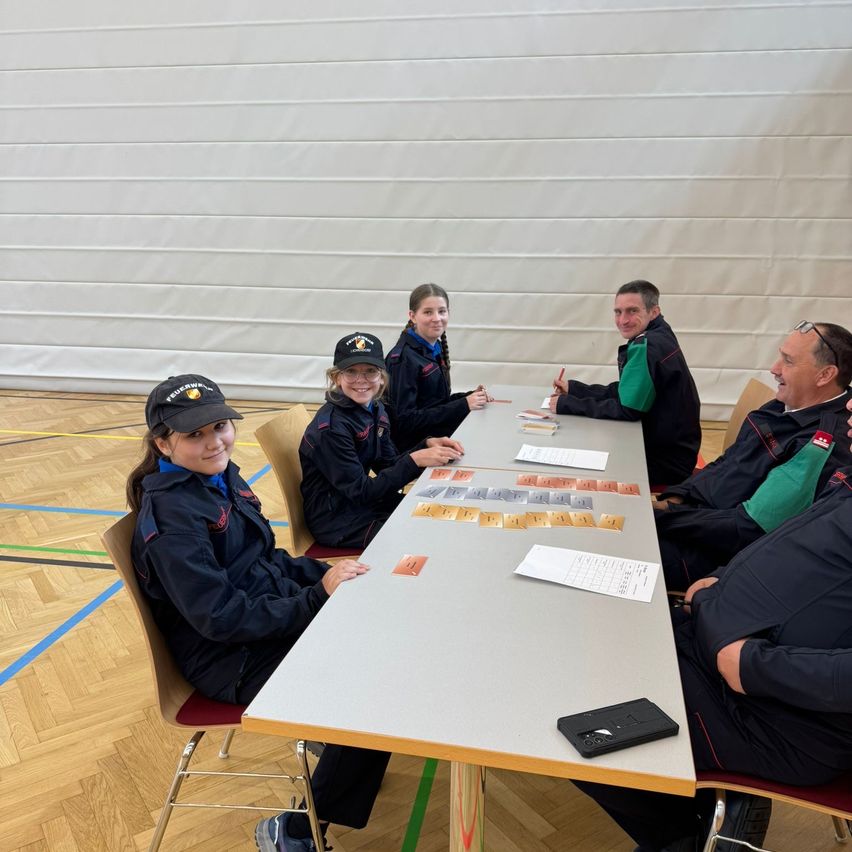 A group of people are sitting around a table in a room. They are wearing uniforms and playing a game. There is a long table with cards, papers, and a notebook.