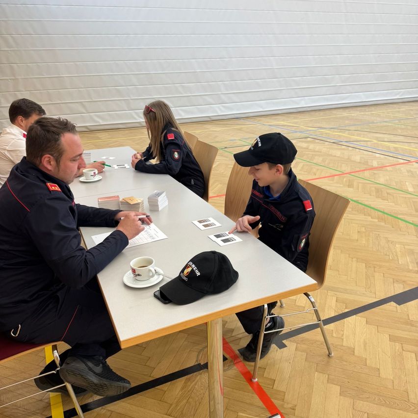 Several individuals are seated around a table in a gym. A man is writing, and a boy looks at him. Two cups and a hat are on the table. A woman sits behind them.
