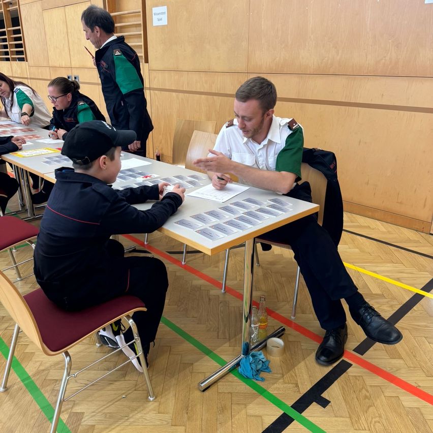 Several individuals are participating in a card game at a table inside a gym. A man is writing on a paper with a pen.