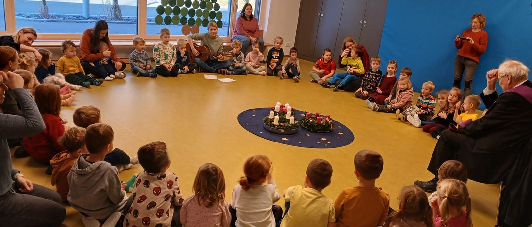 Kinder sitzen im Kreis um einen geschmückten Tisch, während ein Erwachsener Gitarre spielt. Einige Kinder sitzen, andere stehen, und einige Erwachsene sind anwesend. Der Raum hat große Fenster und eine Weihnachtsbaumdekoration.