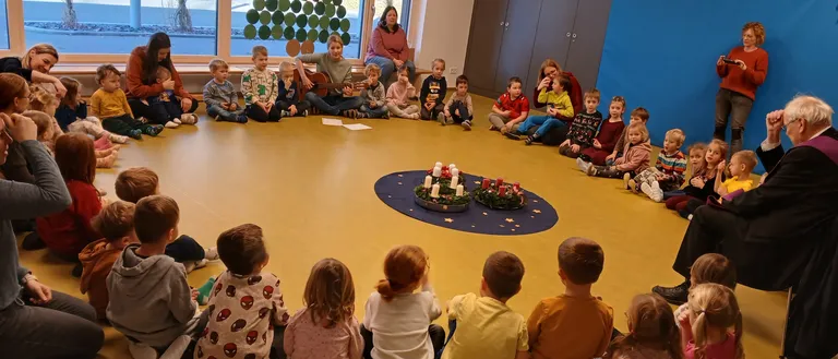 Kinder sitzen im Kreis um einen geschmückten Tisch, während ein Erwachsener Gitarre spielt. Einige Kinder sitzen, andere stehen, und einige Erwachsene sind anwesend. Der Raum hat große Fenster und eine Weihnachtsbaumdekoration.