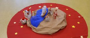 Eine runde rote Matte zeigt eine Miniatur-Krippenszene mit einem Boot, dem Weihnachtsmann, einem Haus und einer Maus, vor dem Hintergrund gelber Sterne.