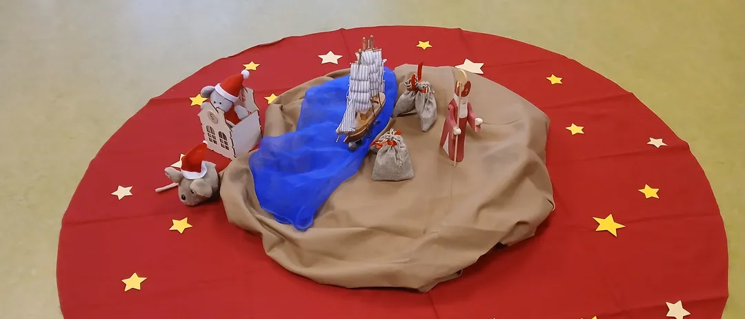 Eine runde rote Matte zeigt eine Miniatur-Krippenszene mit einem Boot, dem Weihnachtsmann, einem Haus und einer Maus, vor dem Hintergrund gelber Sterne.