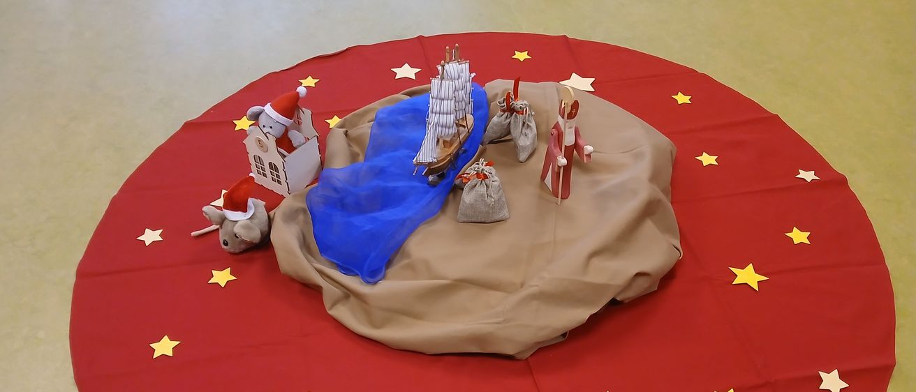 Eine runde rote Matte zeigt eine Miniatur-Krippenszene mit einem Boot, dem Weihnachtsmann, einem Haus und einer Maus, vor dem Hintergrund gelber Sterne.