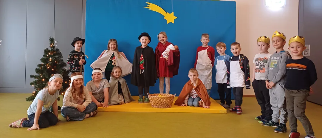 Eine Gruppe von Kindern in verschiedenen Kostümen posiert für ein Foto in einem dekorierten Raum, mit einem blauen Hintergrund, der einen Stern und einen Weihnachtsbaum im Hintergrund zeigt.