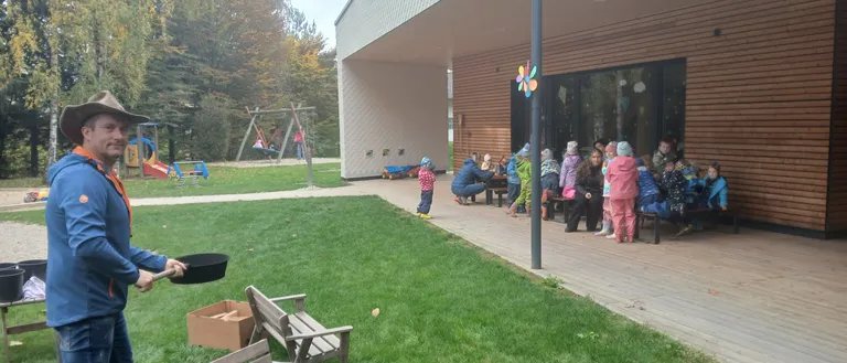 Ein Mann hält eine Pfanne in einem Park. Kinder sitzen auf Bänken und Tischen vor einem Gebäude. Bäume und ein Spielplatz sind im Hintergrund.