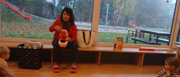 Eine Frau unterrichtet Kinder über Zähne mit einem Modell. Die Kinder sitzen auf dem Boden und hören aufmerksam zu. Durch die großen Fenster ist ein Spielplatz sichtbar.
