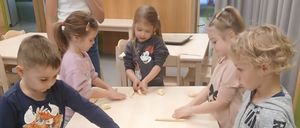 Mehrere Kinder sind um einen Tisch versammelt und beschäftigen sich mit einem Teig. Sie stellen lange, dünne Teigstränge her. Eine Frau steht hinter ihnen und beaufsichtigt die Aktivität.