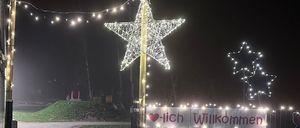 Eine Nachtaufnahme einer Sternendekoration mit Lichtern und einem Banner mit der Aufschrift 'Herzlich Willkommen'. Sie ist beleuchtet und in der Nähe eines Spielplatzes aufgestellt.