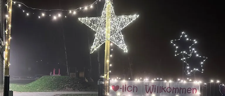 Eine Nachtaufnahme einer Sternendekoration mit Lichtern und einem Banner mit der Aufschrift 'Herzlich Willkommen'. Sie ist beleuchtet und in der Nähe eines Spielplatzes aufgestellt.