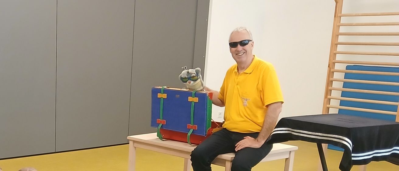 Ein Mann in einem gelben Shirt sitzt auf einer Bank und hält eine Puppe. Kinder sitzen vor ihm auf dem Boden und lauschen.