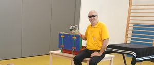 A man in a yellow shirt is sitting on a bench and holding a puppet. Children are sitting on the floor in front of him, listening.