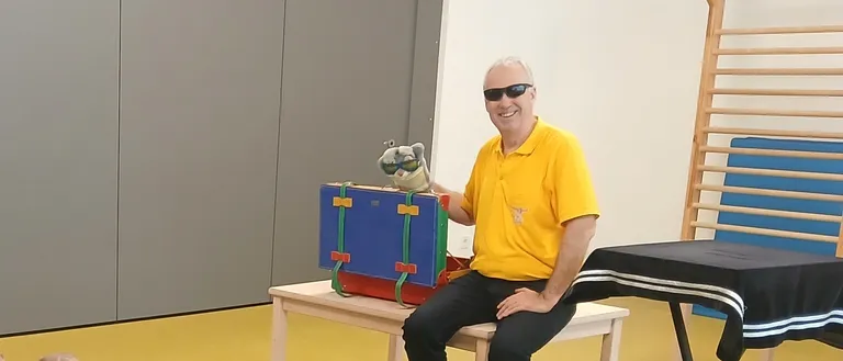 Ein Mann in einem gelben Shirt sitzt auf einer Bank und hält eine Puppe. Kinder sitzen vor ihm auf dem Boden und lauschen.