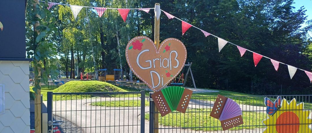 Ein herzförmiges Schild mit der Aufschrift 'GriaB Di' steht neben einem Tor, geschmückt mit Wimpelketten und Akkordeons, in einem Park mit Bäumen und Spielgeräten.