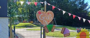 Ein herzförmiges Schild mit der Aufschrift 'GriaB Di' steht neben einem Tor, geschmückt mit Wimpelketten und Akkordeons, in einem Park mit Bäumen und Spielgeräten.