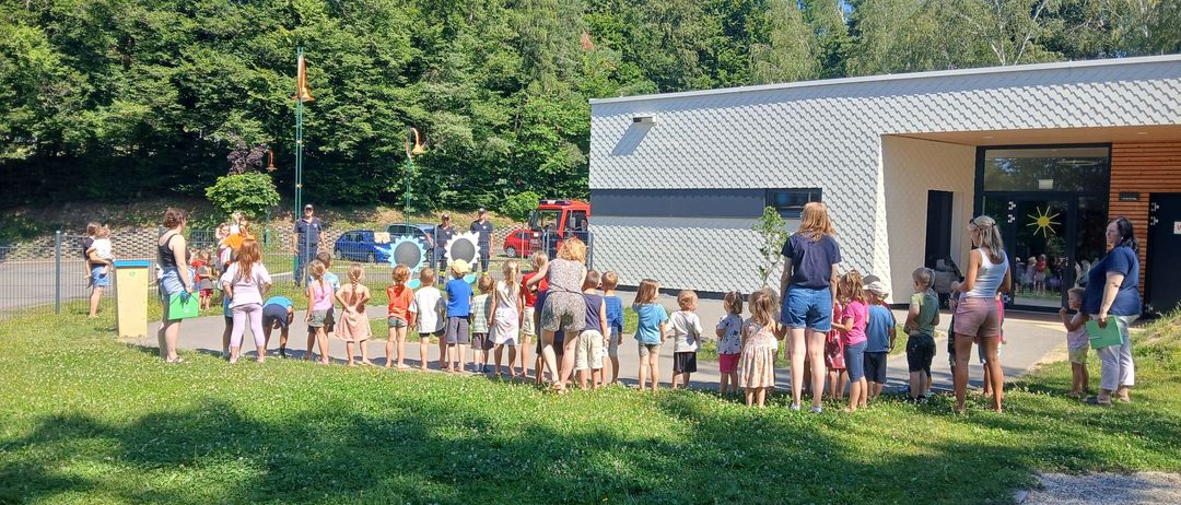 Eine Gruppe von Kindern und Erwachsenen steht in einer Reihe auf einer Wiese. Dahinter ist ein weißes Gebäude mit Glastüren und Fenstern zu sehen. Die Umgebung umfasst mehrere Bäume und geparkte Autos.