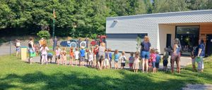 Eine Gruppe von Kindern und Erwachsenen steht in einer Reihe auf einer Wiese. Dahinter ist ein weißes Gebäude mit Glastüren und Fenstern zu sehen. Die Umgebung umfasst mehrere Bäume und geparkte Autos.