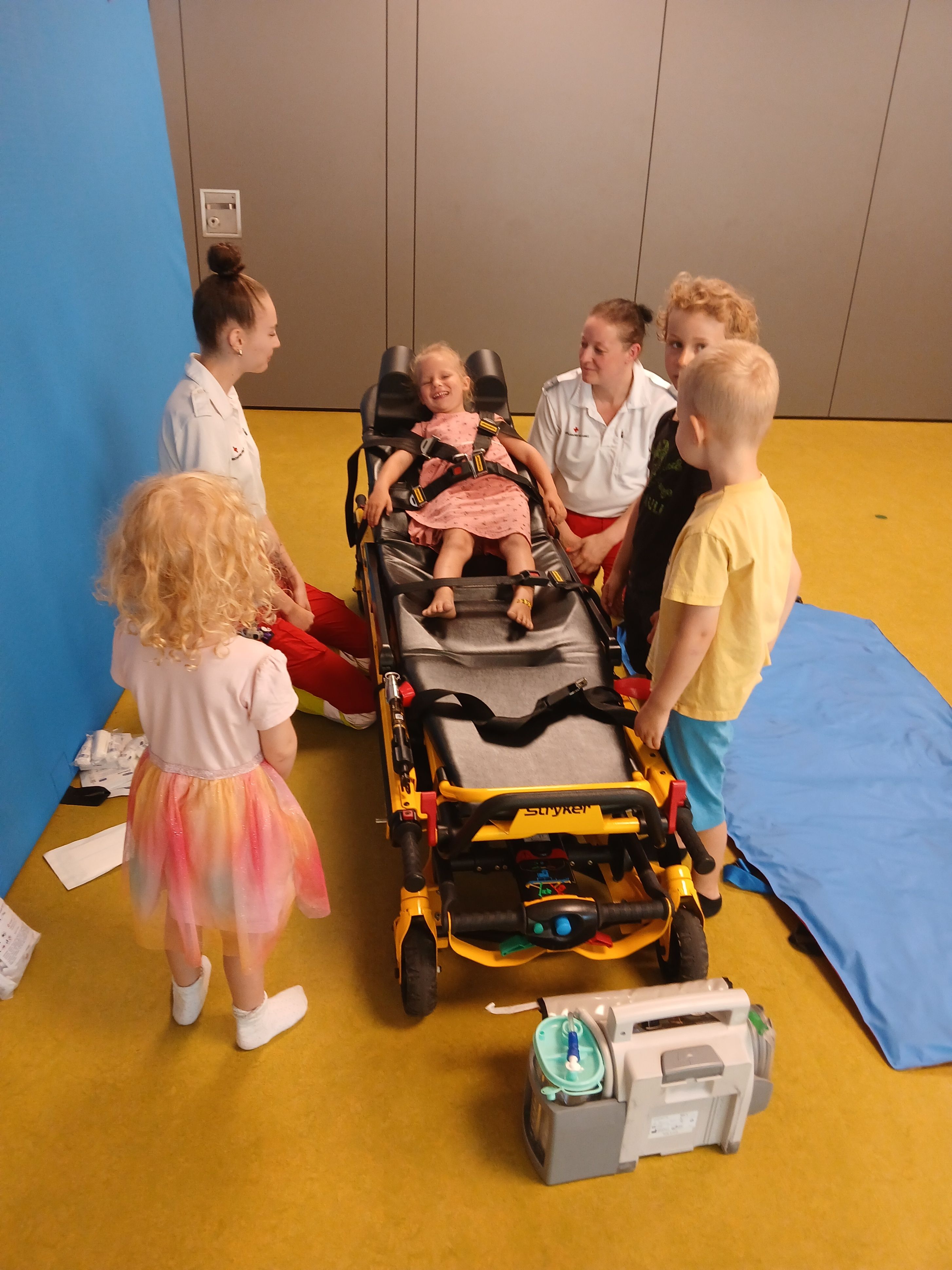 Eine Gruppe von Kindern ist um einen Krankenwagen versammelt. Ein kleines Mädchen sitzt auf dem Krankenwagen, gesichert mit einem Gurt, während zwei Erwachsene sich um sie kümmern. Andere Kinder stehen in der Nähe und beobachten. Die Umgebung scheint ein Simulationsraum zu sein.