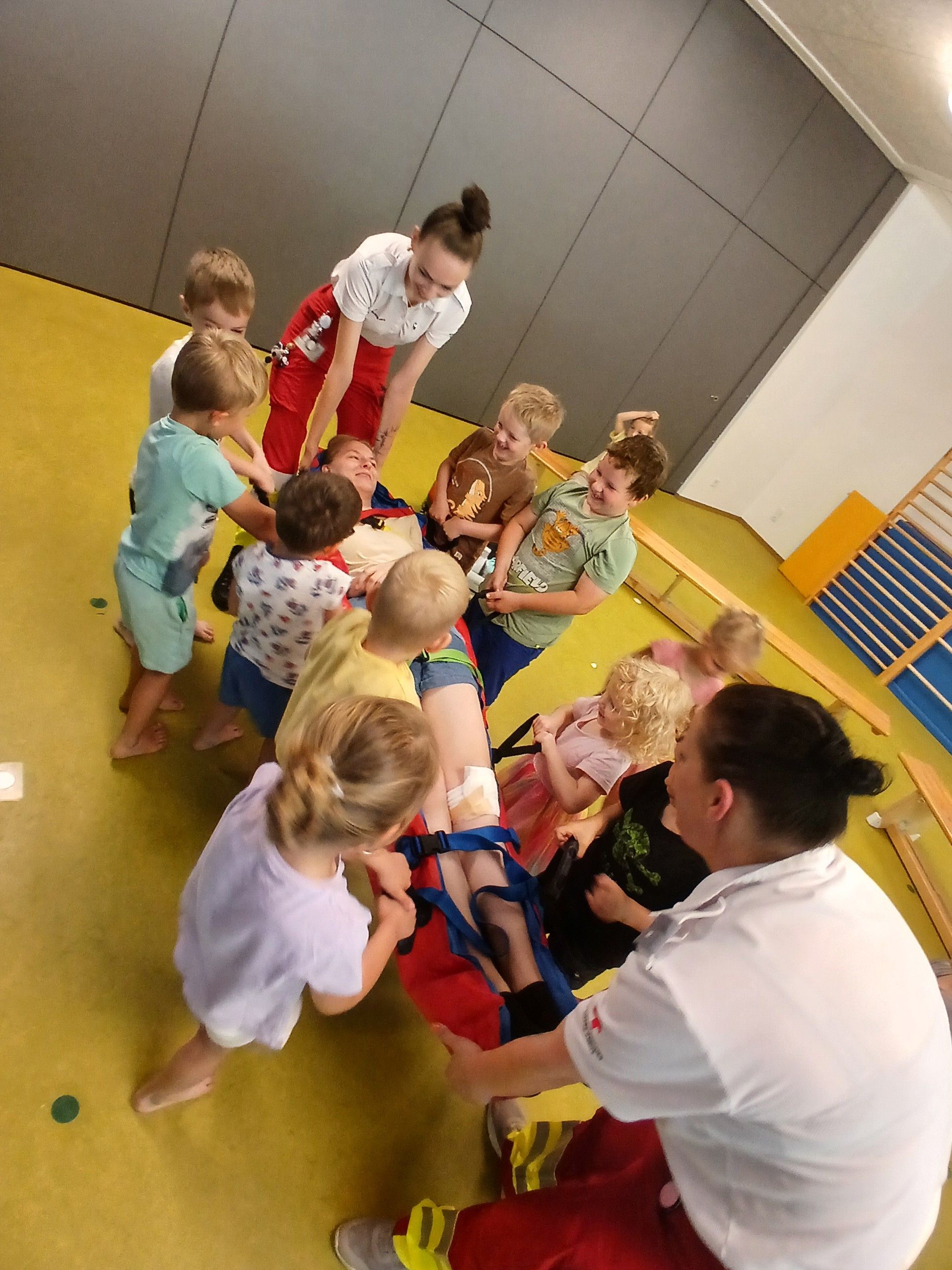 Eine Gruppe von Kindern befindet sich in einem Fitnessstudio mit einer Frau, die ein Kind in einem Gurtzeug hält. Die Frau hilft dem Kind, aufrecht zu stehen.