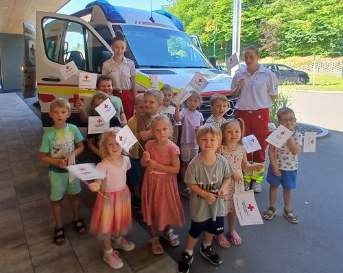 Eine Gruppe von Kindern und zwei Erwachsenen halten weiße Flaggen mit roten Kreuzen und stehen vor einem Krankenwagen. Sie lächeln und scheinen für ein Foto zu posieren.