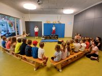 Zwei Personen in roten und weißen Uniformen demonstrieren Notfallausrüstung in einem Klassenzimmer. Kinder sitzen auf Bänken und beobachten aufmerksam. Ein blauer Hintergrund mit Cartoon-Charakteren steht hinter den Präsentatoren.