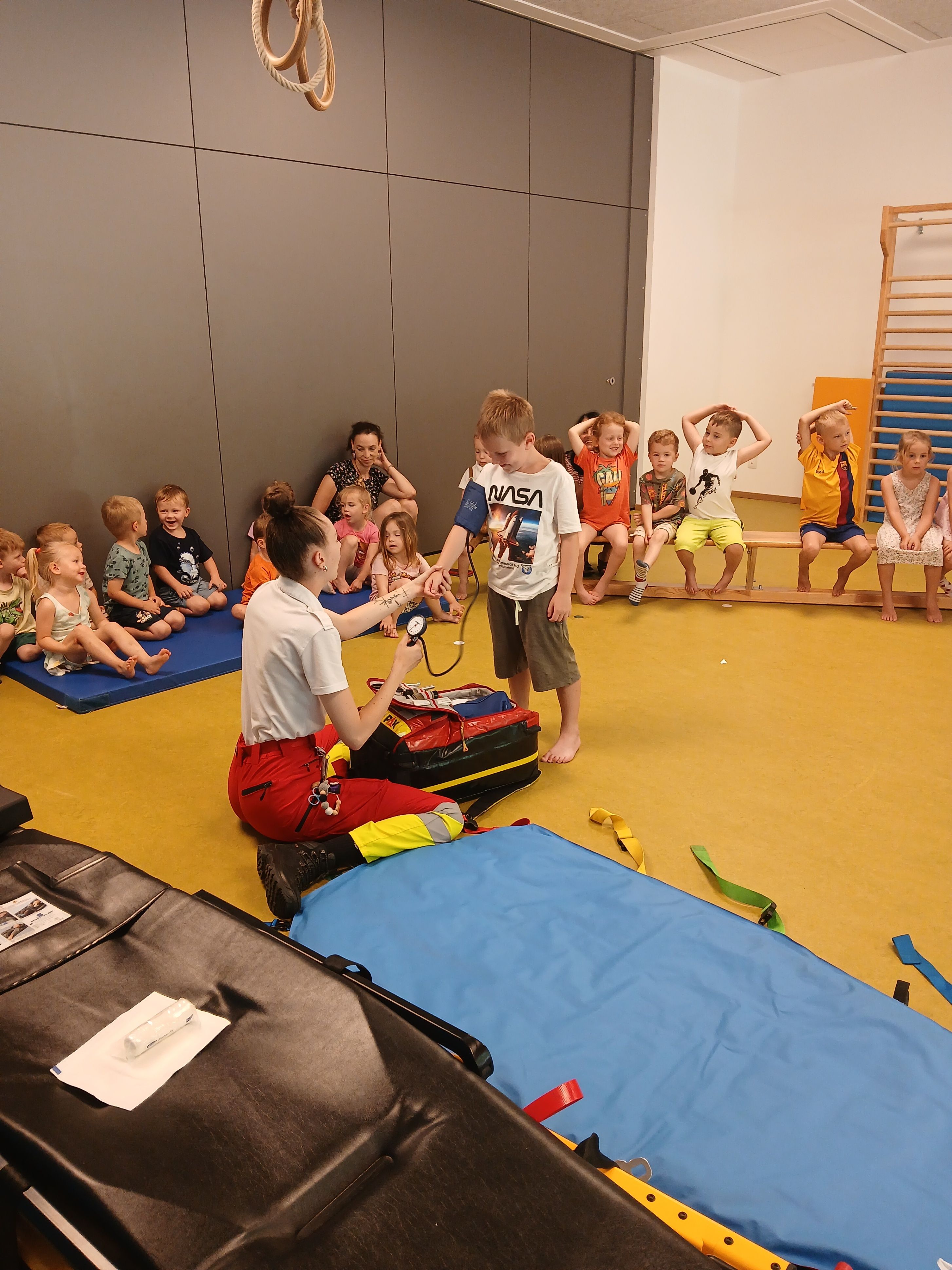 Eine Krankenschwester misst den Blutdruck eines Jungen, während andere zuschauen. Mehrere Kinder sitzen auf dem Boden, während andere auf einer Bank sitzen. Eine Holzleiter und Matten sind in der Nähe.