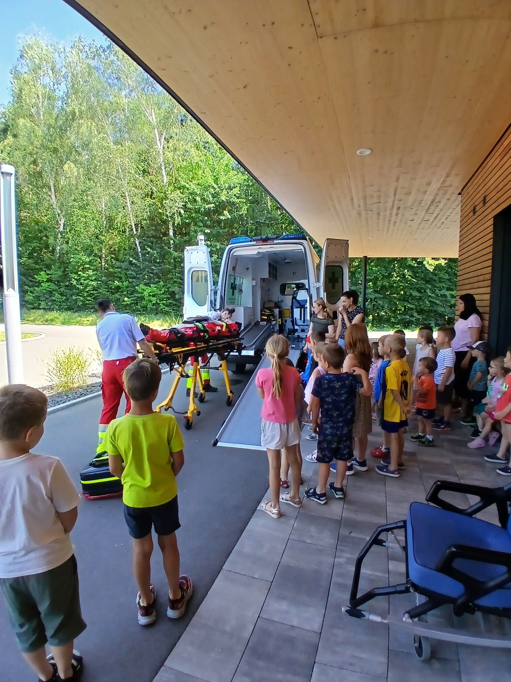 Ein Sanitäter transportiert einen Patienten auf einer Trage von einem Krankenwagen. Kinder stehen draußen und beobachten die Szene. Der Krankenwagen ist am Straßenrand geparkt.