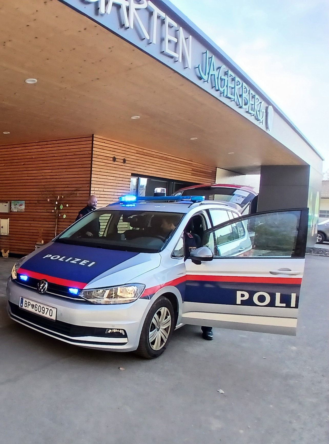 Ein Polizeiauto ist vor einem Gebäude geparkt, mit dem Wort 'POLIZEI' an der Seite und auf dem Dach. Die Autotür steht offen, und ein Polizist steht in der Nähe. Das Gebäude hat Holzwände und ein Schild mit der Aufschrift 'JAGERBERG.'