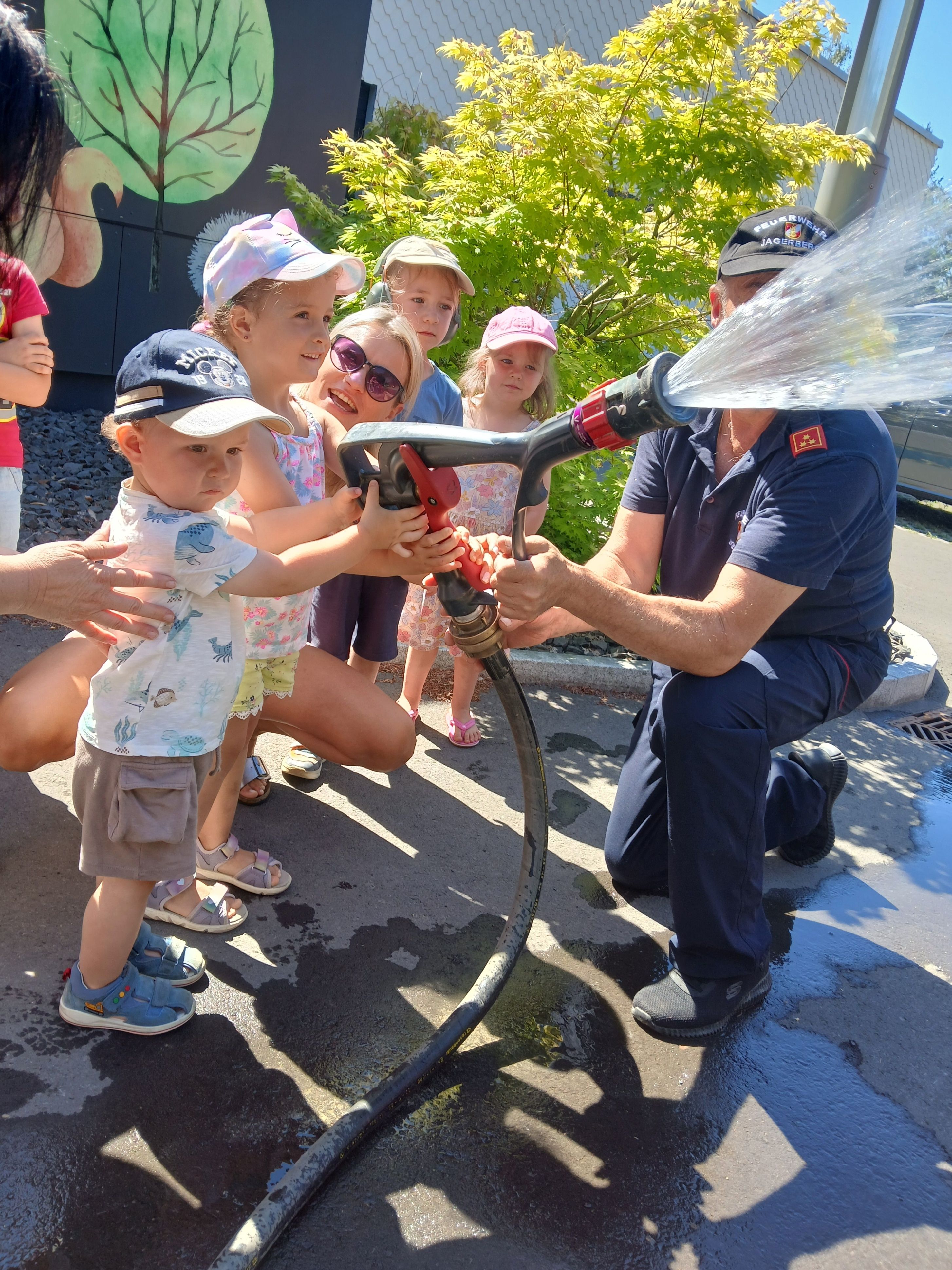Ein Feuerwehrmann hilft Kindern, einen Feuerwehrschlauch zu halten und mit dem Wasser zu spielen. Fünf Kinder und ein Erwachsener sind draußen.