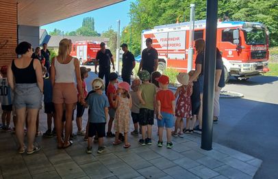 Eine Gruppe von Kindern und Erwachsenen steht draußen, mit drei Feuerwehrleuten vor ihnen. Sie sind in der Nähe eines Feuerwehrwagens mit dem Wort 'Feuerwehr' darauf. Im Hintergrund sind zwei weitere Feuerwehrwagen.