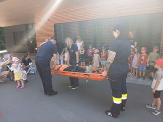 Zwei Rettungssanitäter tragen eine orangefarbene Trage mit einer Person davor vor einer Menge von Kindern.