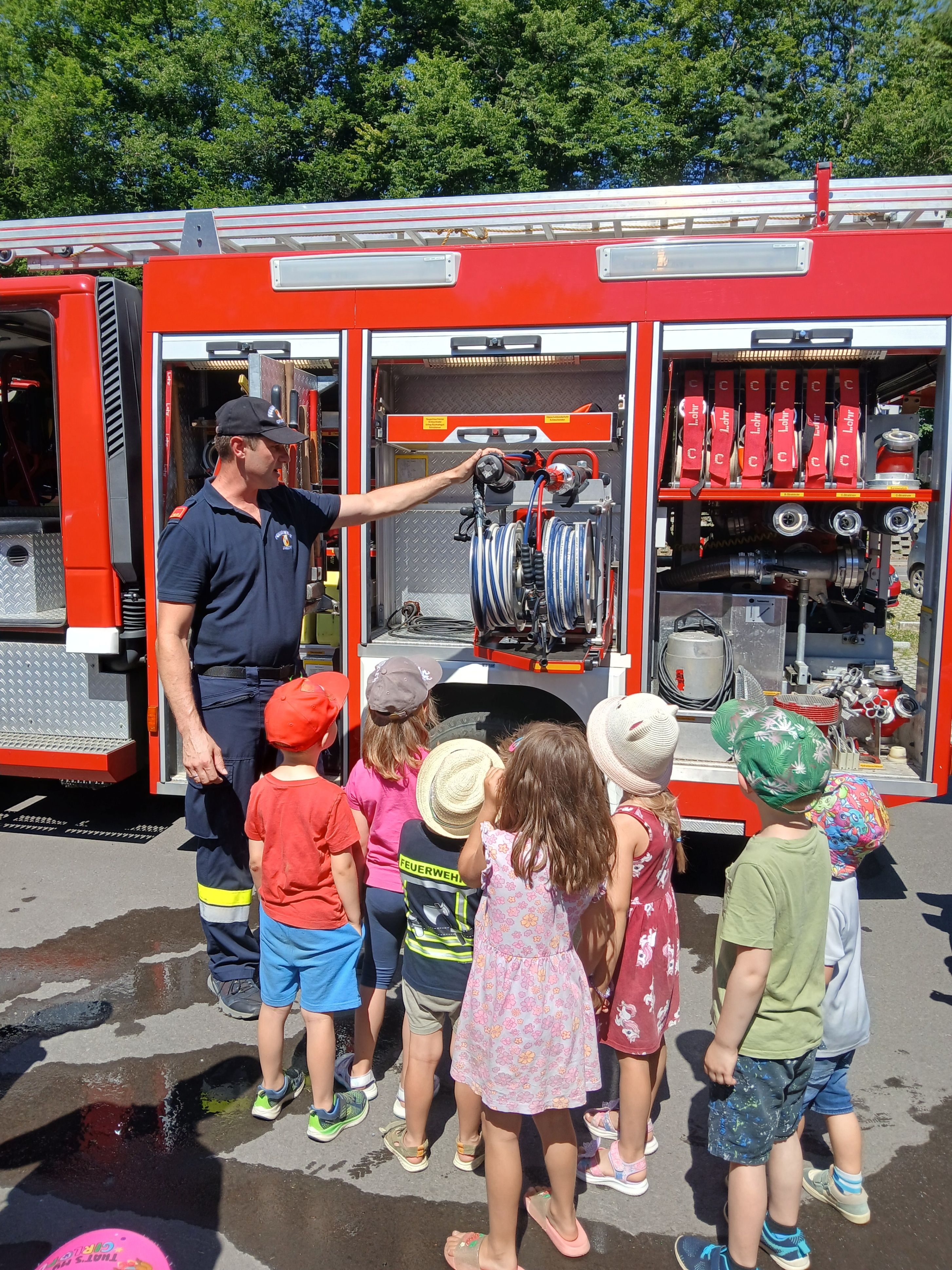 Ein Feuerwehrmann zeigt einer Gruppe von Kindern, wie das Feuerwehrauto funktioniert. Die Kinder tragen Mützen und Turnschuhe und stehen neben dem Feuerwehrauto.