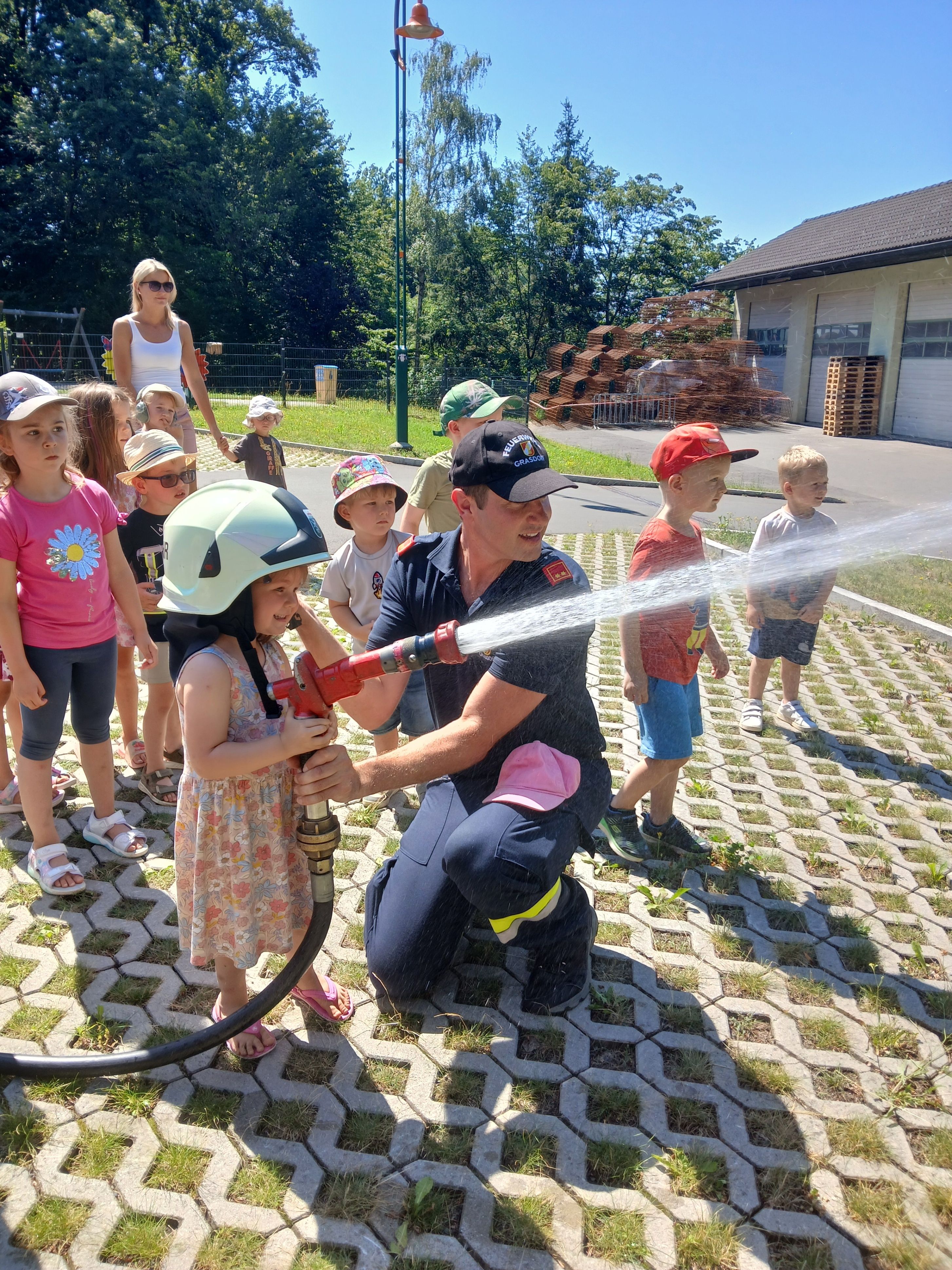 Ein Feuerwehrmann zeigt Kindern, wie man einen Feuerwehrschlauch benutzt, während eine Frau hinter ihnen steht. Ein Gebäude und ein Spielplatz sind im Hintergrund zu sehen.