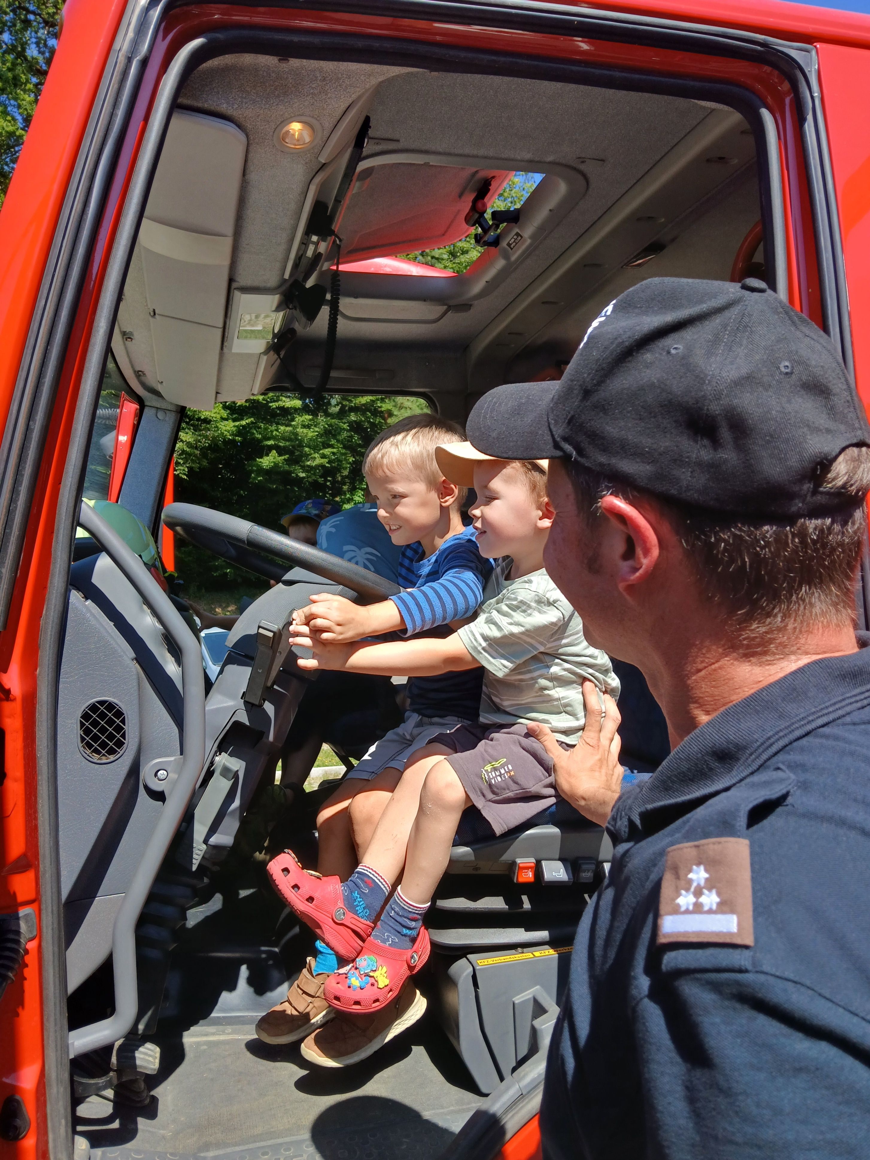 Zwei Jungen sitzen auf dem Fahrersitz eines roten Feuerwehrwagens, einer hält das Lenkrad, während ein Mann in Uniform aufmerksam zusieht.