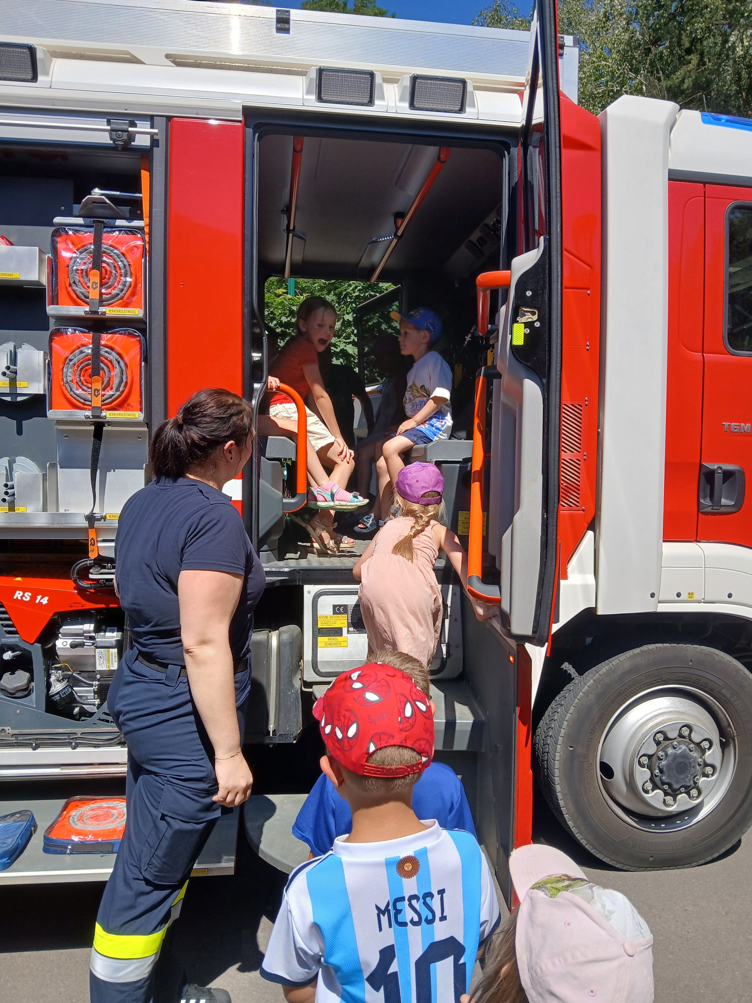Ein Feuerwehrmann zeigt einer Gruppe von Kindern einen Feuerwehrwagen. Vier Kinder sind im Wagen, während eine Frau draußen steht.