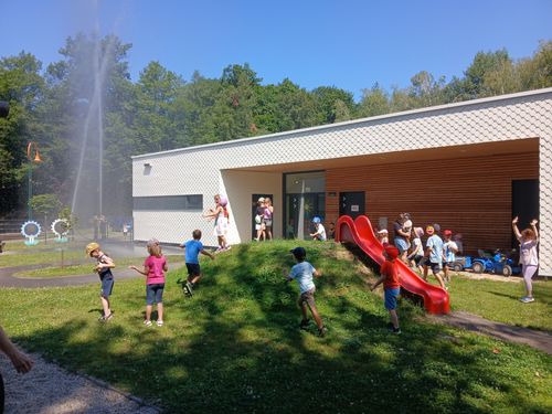 Kinder spielen vor einem Gebäude mit einer roten Rutsche. Eine Fontäne sprüht im Vordergrund Wasser.