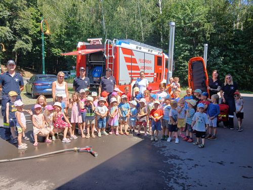 Eine Gruppe von Kindern und Erwachsenen posiert vor einem roten Feuerwehrauto, mit einem Feuerhydranten und Bäumen im Hintergrund.