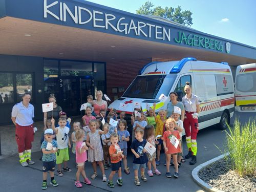 Eine Gruppe von Kindern und Erwachsenen versammelt sich vor einem Kindergarten Jagerberg-Gebäude. Sie halten kleine Flaggen und lächeln in die Kamera. Ein Krankenwagen ist in der Nähe geparkt.