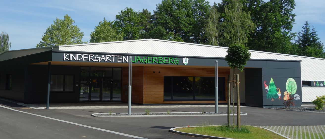 Das Äußere des Kindergartens Jagerberg ist zu sehen, mit einem modernen Gebäude, großen Fenstern, einem Baum davor und einem klaren blauen Himmel mit verstreuten Wolken.