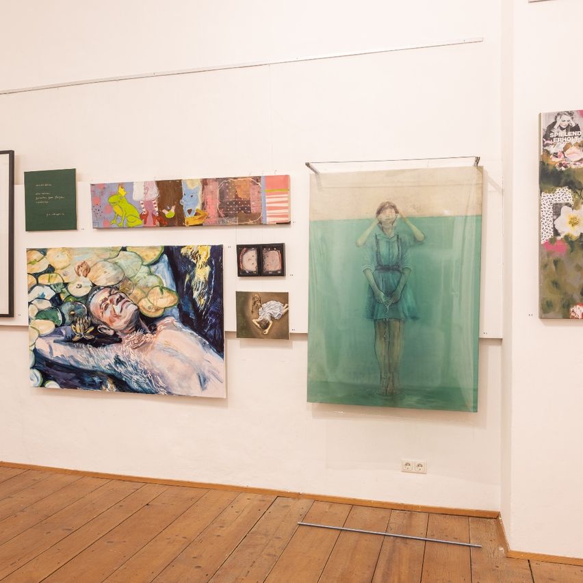 Ein Kunstgalerie mit weißen Wänden zeigt verschiedene Gemälde, darunter eine Frau in einem blauen Kleid, die im Wasser steht. Andere Gemälde zeigen abstrakte Designs und eine liegende Person.