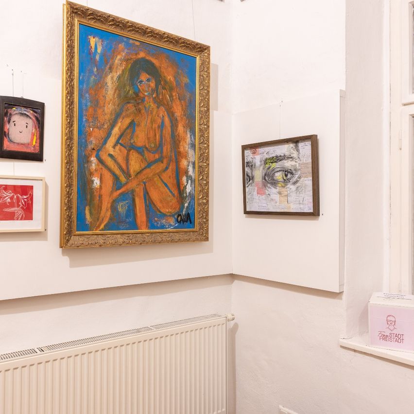 Ein Raum mit weißer Wand, der verschiedene Kunstwerke zeigt. Ein großes gerahmtes Gemälde einer Frau befindet sich mittig. Links hängen kleinere gerahmte Stücke. Unten steht ein weißer Heizkörper und ein Karton mit der Aufschrift 'Die Stadt' auf der Fensterbank.