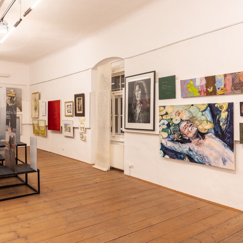 Ein Kunstgalerie mit verschiedenen Gemälden an der Wand. Mehrere Kunstwerke sind in Rahmen ausgestellt. Ein Holzboden und weiße Wände. Ein Fenster und Heizkörper im Hintergrund.