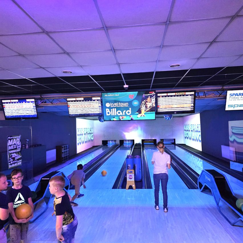 Kids and adults are bowling at a bowling alley with several lanes and monitors displaying scores. A woman is watching the game while others hold bowling balls.