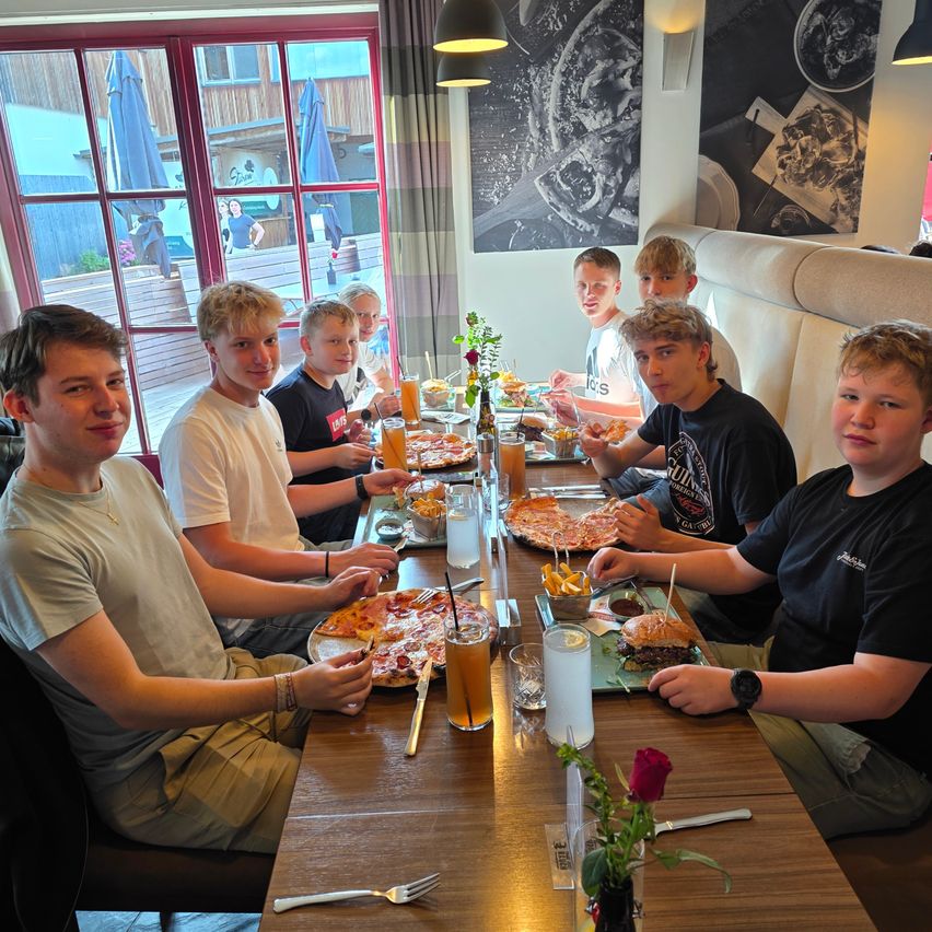 A group of boys are seated around a table in a restaurant, eating pizza and hamburgers. They have drinks and utensils in front of them.