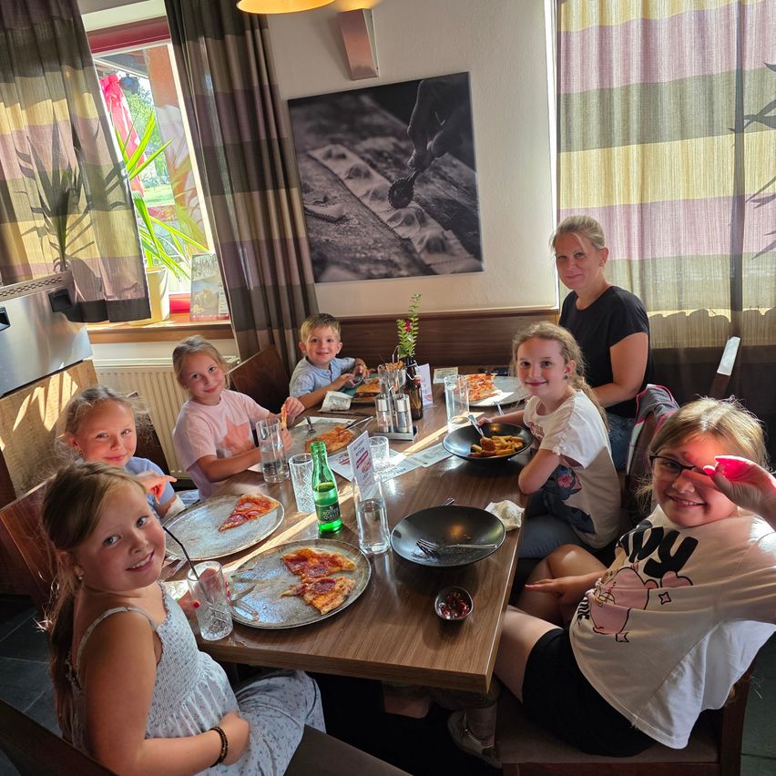 A group of children and adults enjoy a meal at a restaurant. Pizza, glasses, and bowls are on the table. A woman stands behind the children.