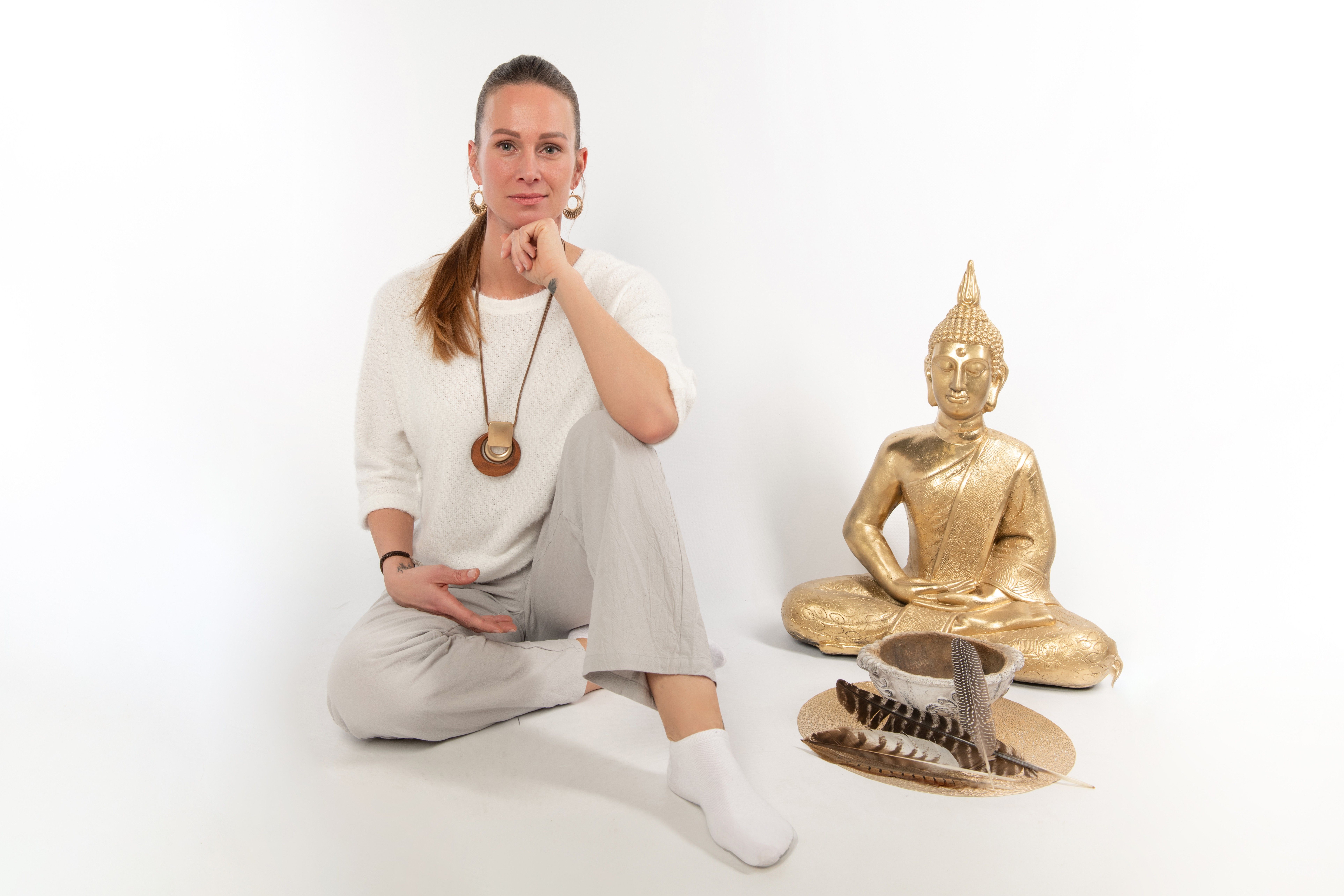 Eine Frau in weißer Kleidung sitzt mit verschränkten Beinen und Kinn in der Hand neben einer goldenen Buddha-Statue und einer Schale mit Federn.