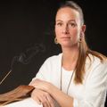Eine Frau in einem weißen Pullover mit langen Haaren sitzt neben einem Räucherstäbchen und Halter und schaut in die Kamera.