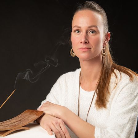 Eine Frau in einem weißen Pullover mit langen Haaren sitzt neben einem Räucherstäbchen und Halter und schaut in die Kamera.