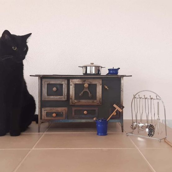 Eine schwarze Katze sitzt neben einem kleinen Ofen mit einem Topf darauf. Der Ofen hat Schubladen und einen Holzgriff. Ein Utensilienständer aus Metall befindet sich in der Nähe.